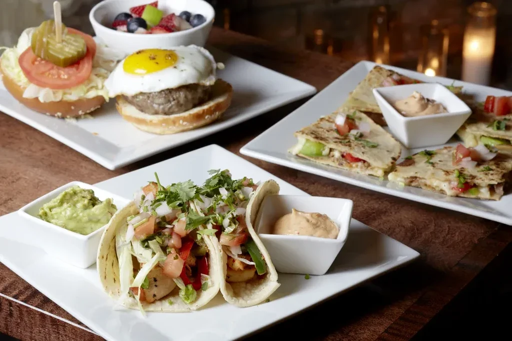 Various dishes including tacos, burgers, quesadillas, and fruit on white plates.