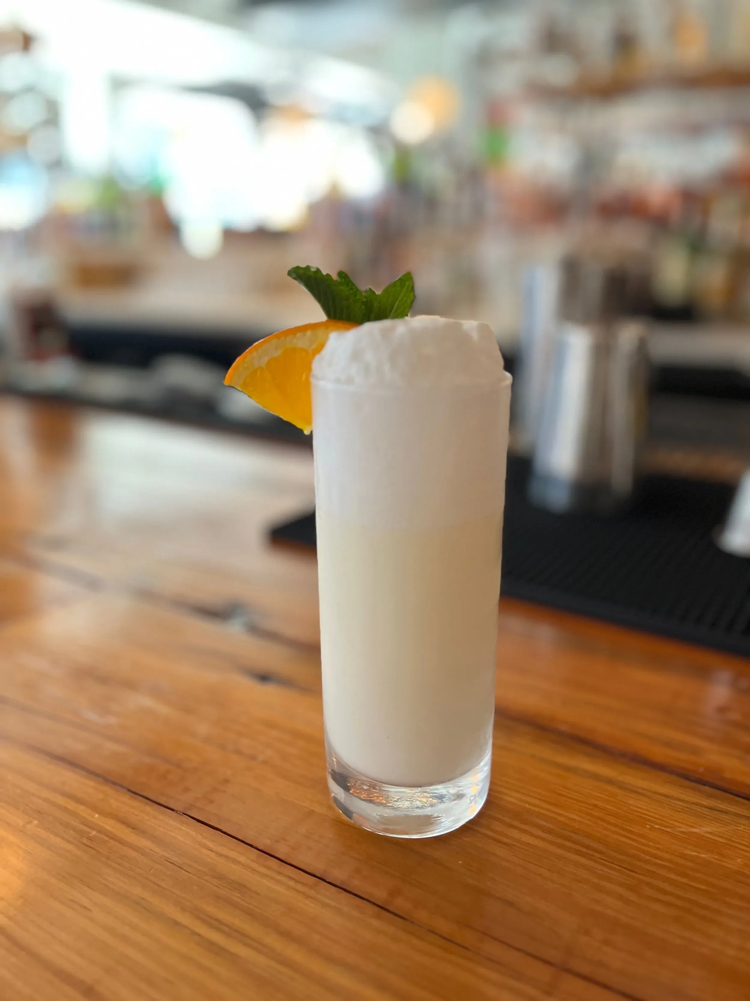 A tall white cocktail with a foamy head, garnished with orange and mint.