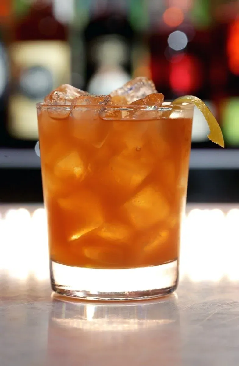 A brown cocktail with ice and a lemon peel on a bar.