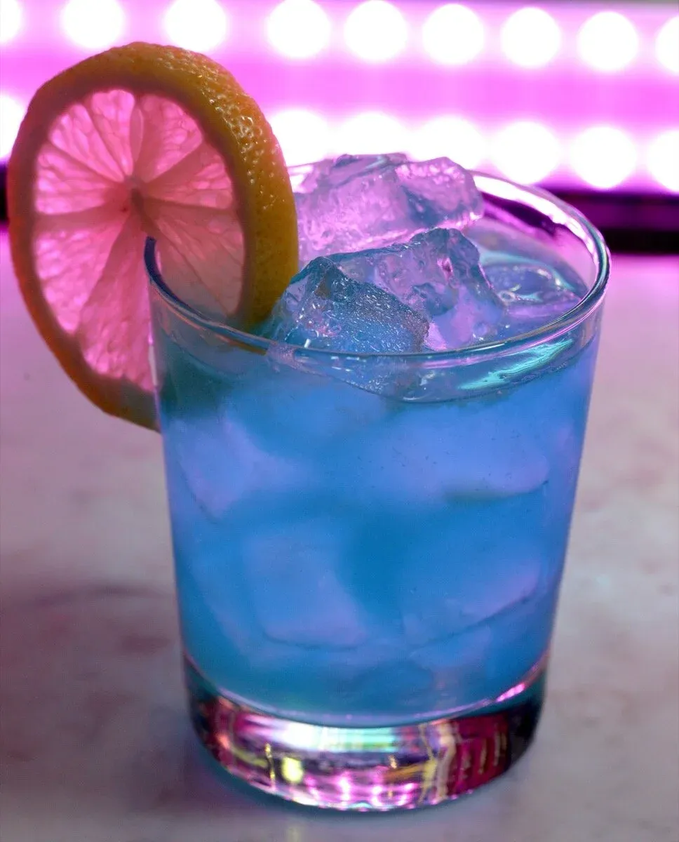 A blue cocktail with ice and a lemon slice, backlit by pink lights.