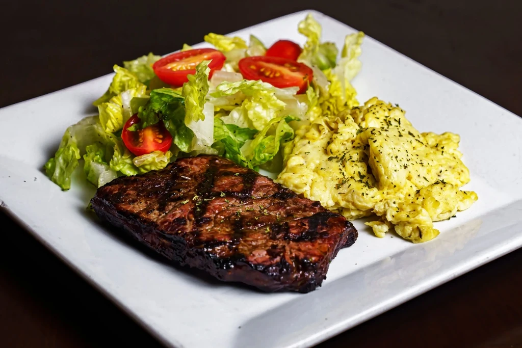 Grilled steak, scrambled eggs, and green salad with cherry tomatoes on a plate.