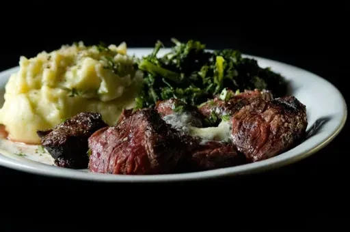 Cooked steak, mashed potatoes, and greens on a white plate.