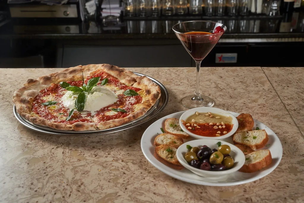 A table with pizza, appetizers, and a cocktail in a restaurant.