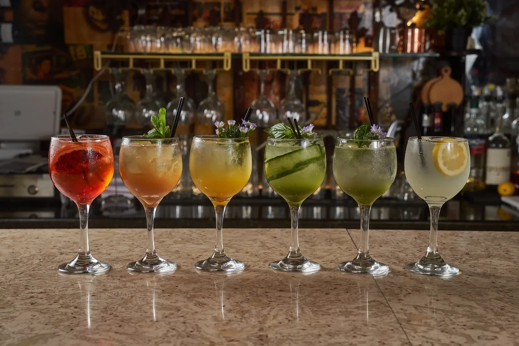 Six colorful cocktails in tall glasses with garnishes on a bar.