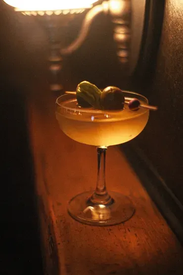 A golden cocktail in a coupe glass with olive and pickled pepper garnish.