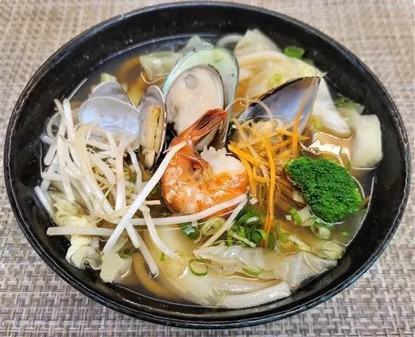 A black bowl of seafood noodle soup with vegetables.