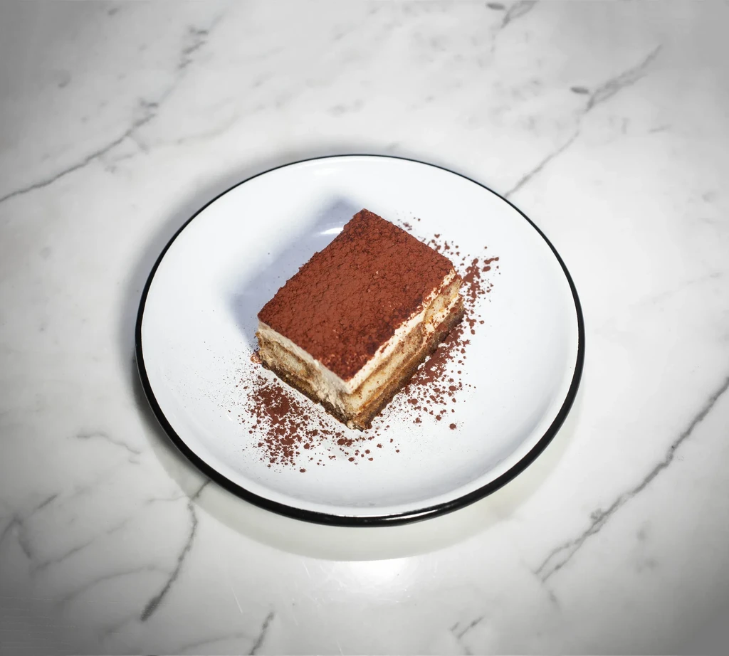 A tiramisu slice dusted with cocoa powder on a white plate.