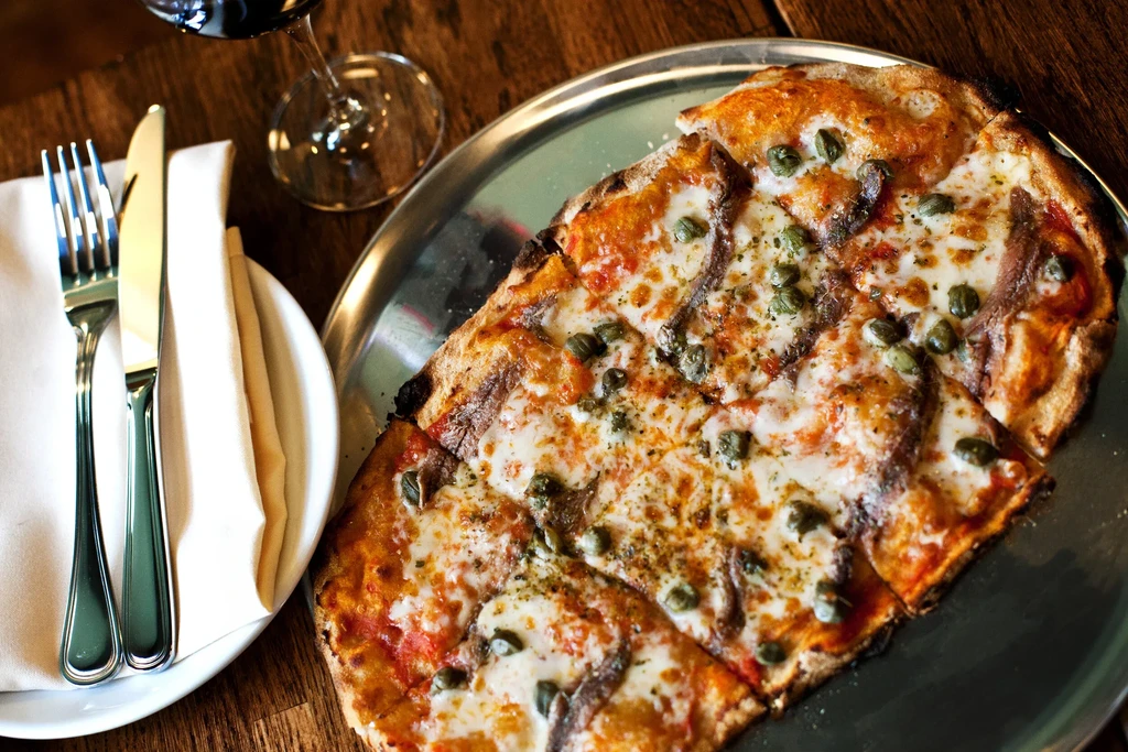 Flatbread pizza with anchovies and capers, cutlery, and wine glass.