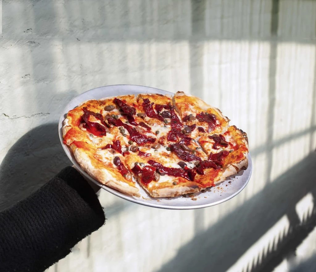 A hand holding a whole pizza with red topping, olives, and capers.