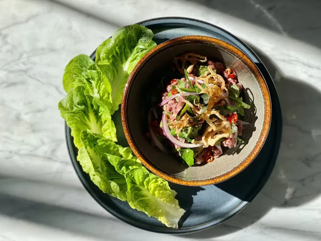A bowl of laap with lettuce wraps on a marble surface.