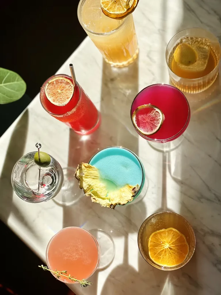 Seven colorful cocktails on a marble bar in bright sunlight.