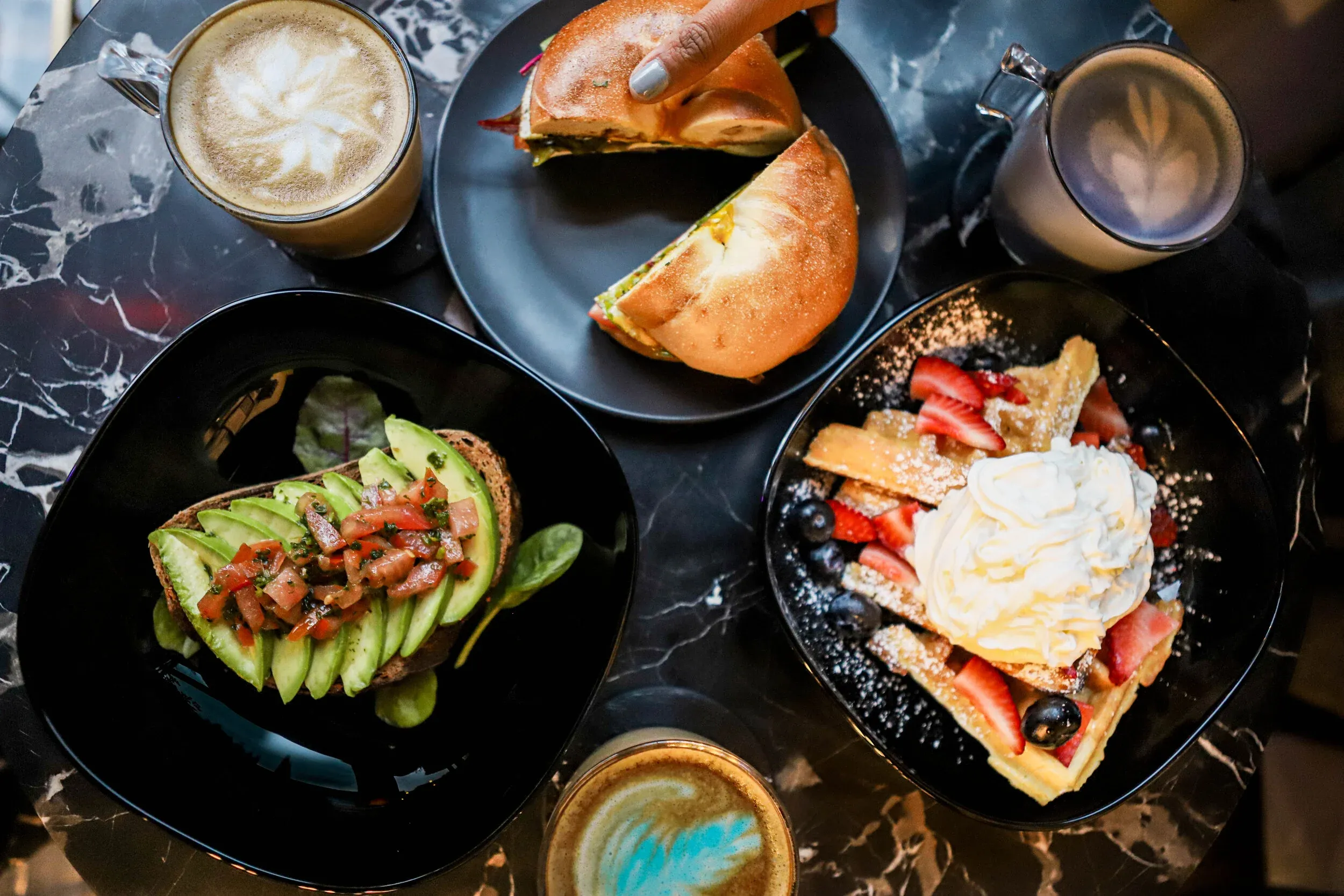 Top-down view of breakfast food: avocado toast, waffles, bagel, and coffee.