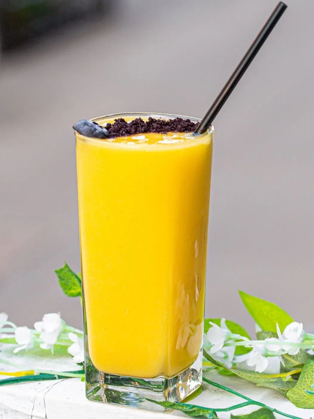 A tall glass of yellow smoothie with dark crumble and a blueberry.