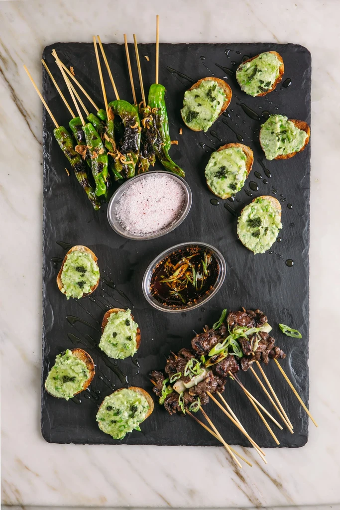 Grilled peppers, meat skewers, green toasts, salt and sauce on slate.