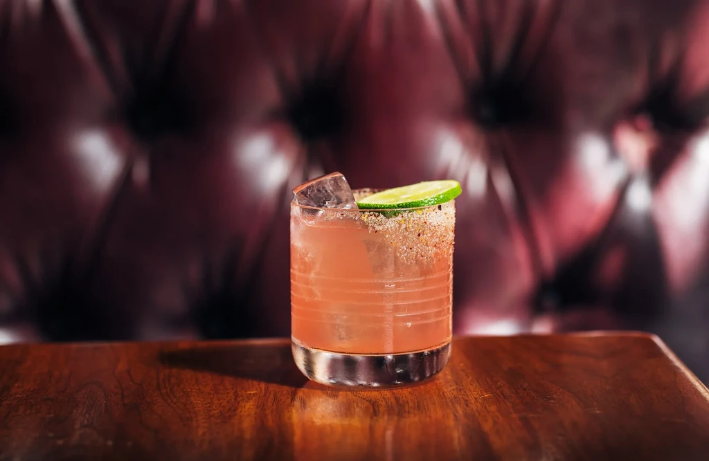 A pink cocktail in a glass with a sugared rim and lime.