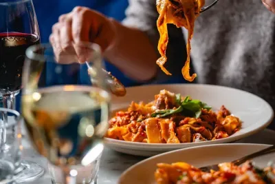 A person eating pasta with a fork, with glasses of wine in the foreground.