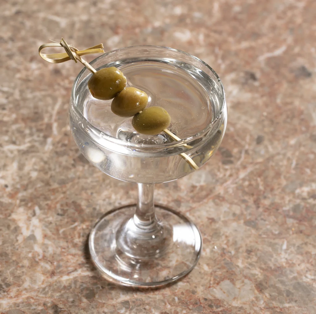 A martini glass with three olives on a skewer on a marble counter.