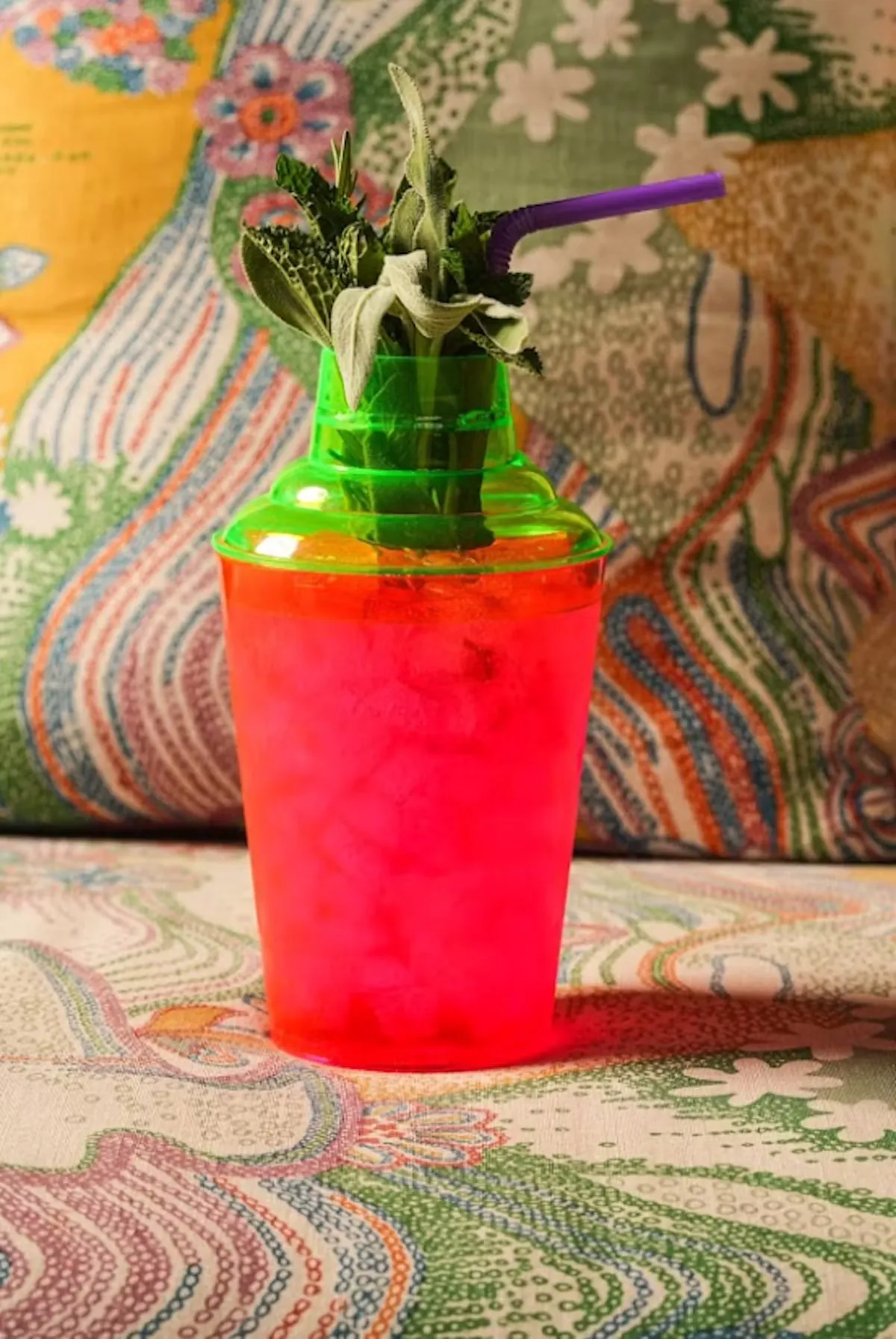 A pink plastic cup with a green lid, fresh herbs, and purple straw.