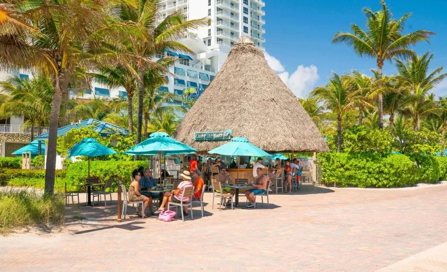 Sunny tropical beach bar with patrons, palm trees, and hotel.