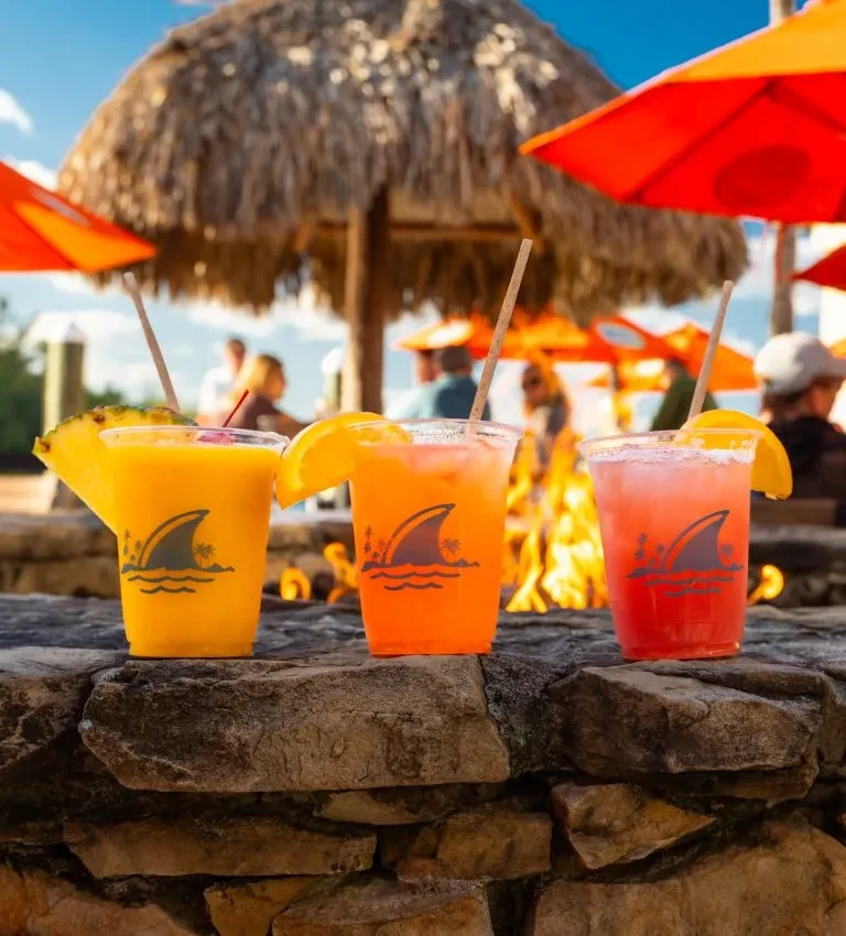 Three vibrant tropical drinks with shark logos at an outdoor resort.