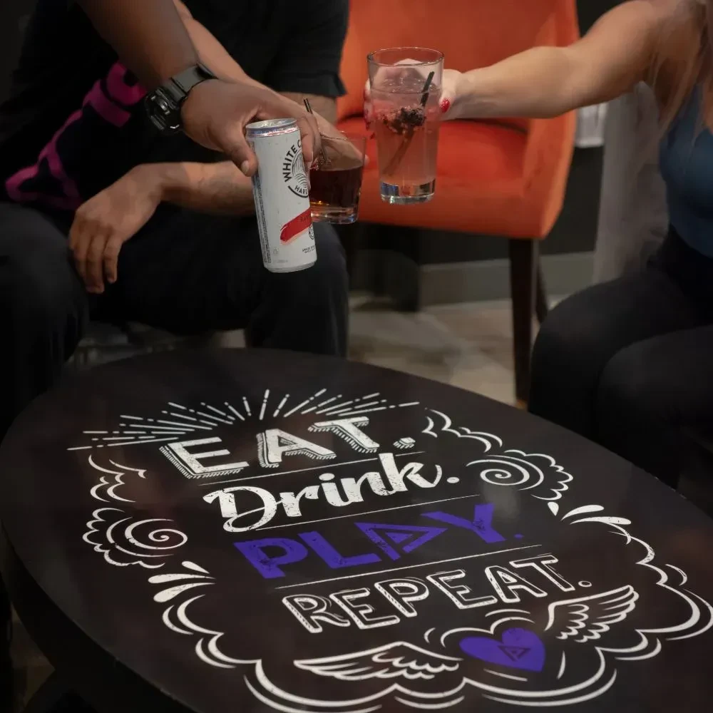 Two people toast drinks over a table with "EAT. Drink. PLAY. REPEAT." text.