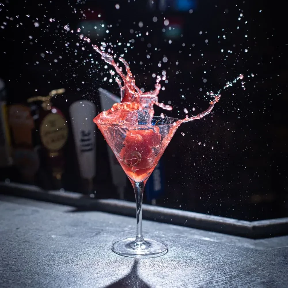 Red cocktail splashes from martini glass with fruit on bar.
