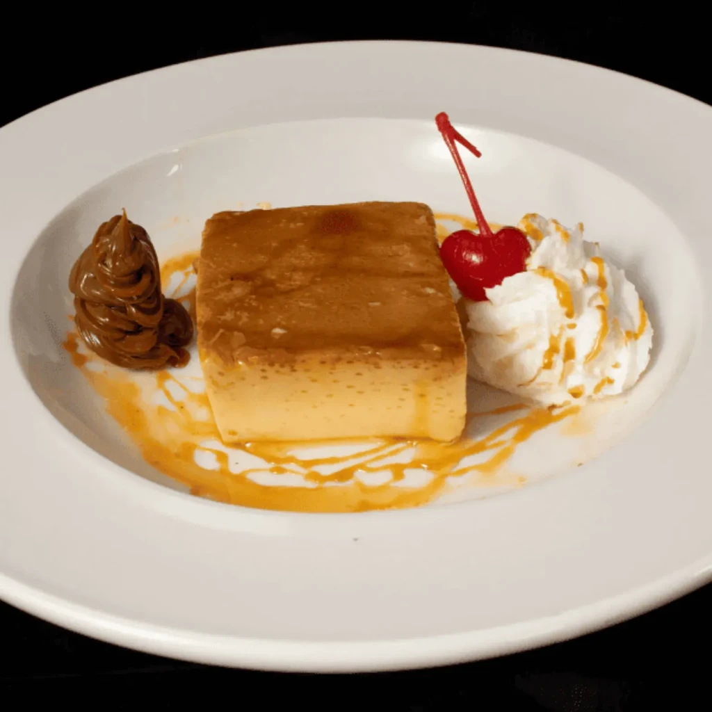 Rectangular flan with caramel, whipped cream, and cherry on white plate.