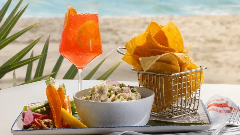 A vibrant cocktail, fresh vegetables, dip, and chips by the beach.