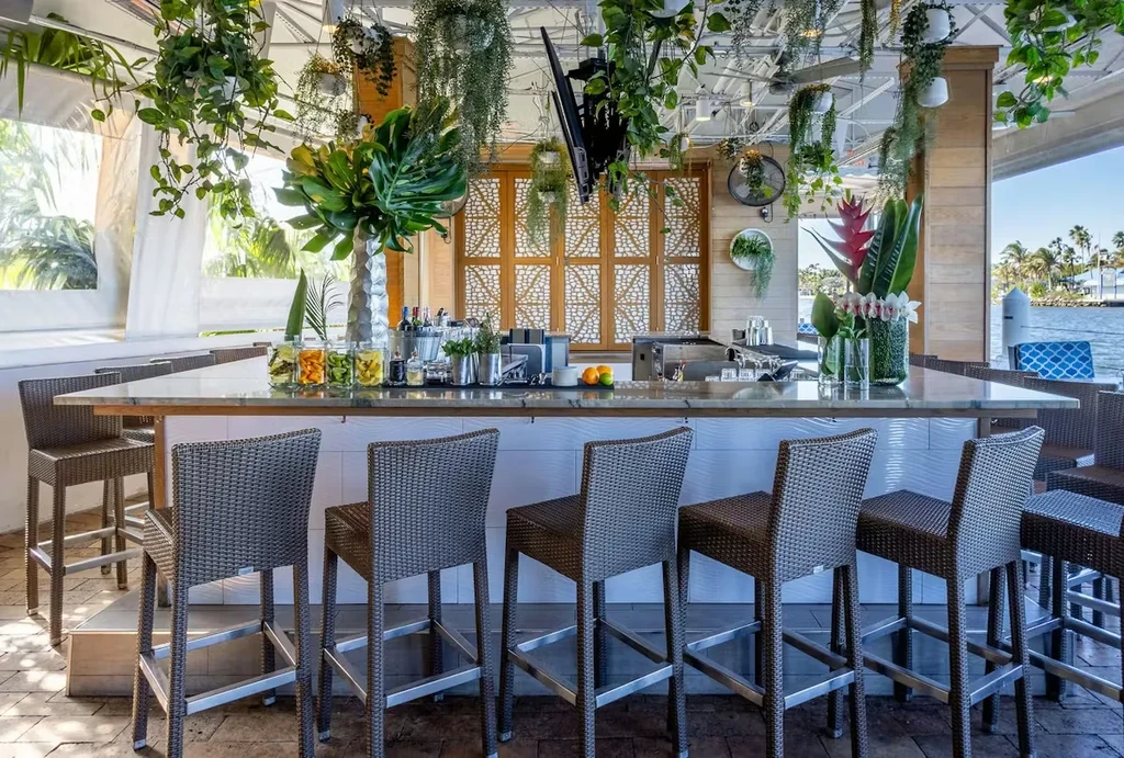 Empty marble bar with woven stools, tropical plants, waterfront setting.