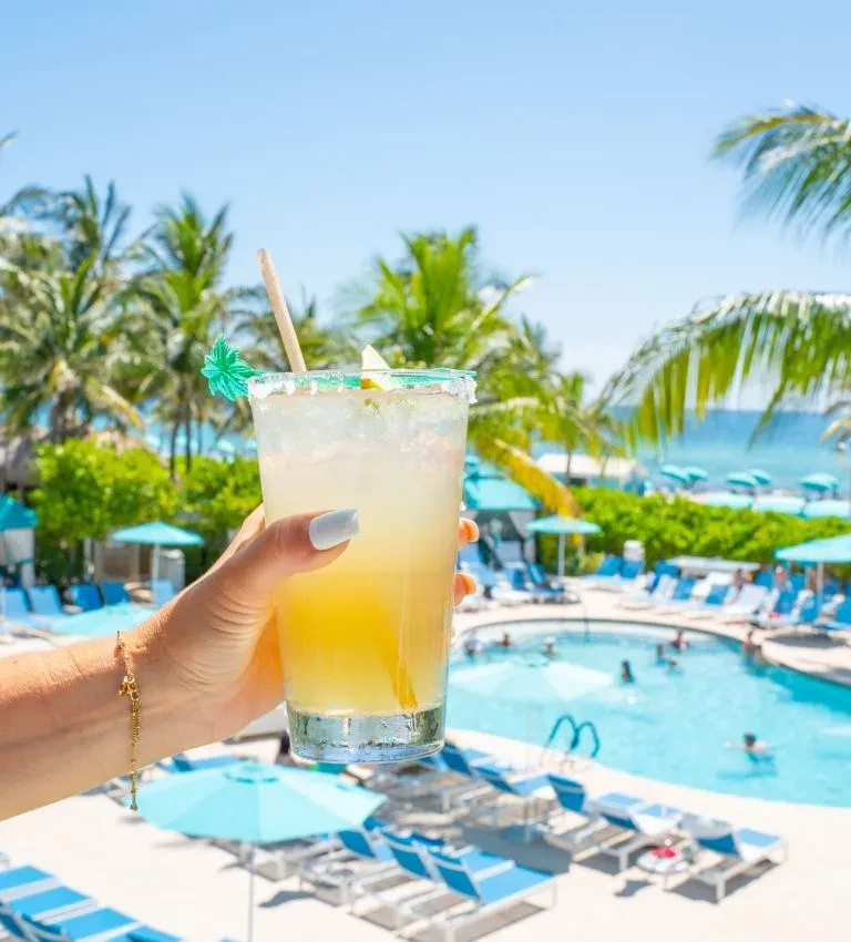 Hand holding a cocktail with lime and umbrella by a sunny pool.