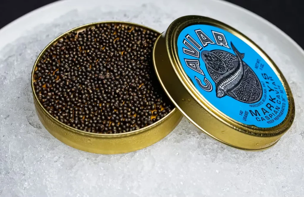 Open golden tin of dark caviar with blue label on crushed ice.