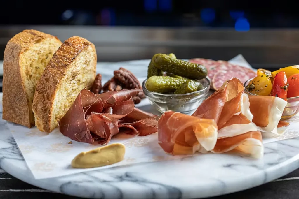 Marble charcuterie board with meats, bread, gherkins, peppers, and nuts.