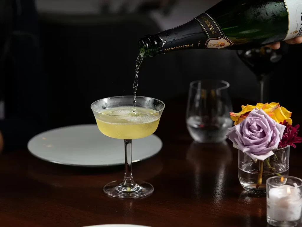 Hand pours golden sparkling wine into coupe glass on dark table.