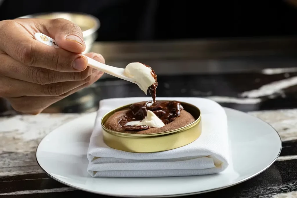 Hands drip cream onto a chocolate dessert in a golden tin.