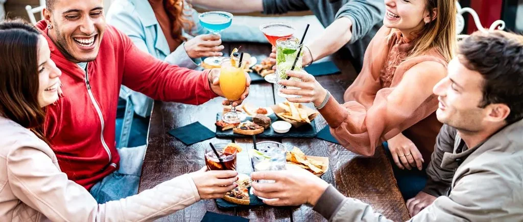 Friends toast colorful cocktails and appetizers at a table.