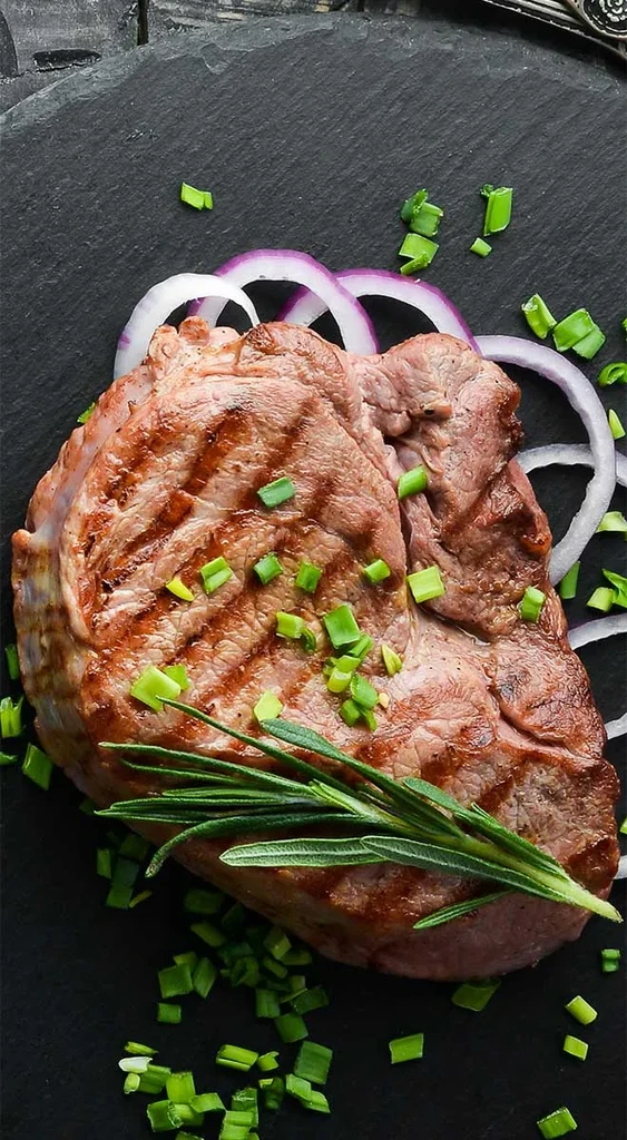 Grilled steak with char marks, rosemary, and green onions, on a dark plate.