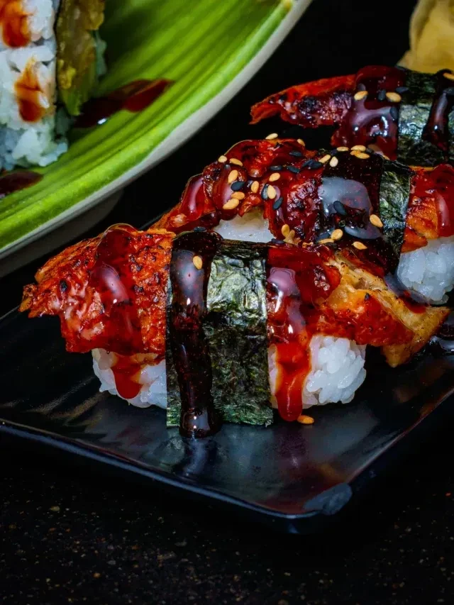 Unagi nigiri with glossy sauce and sesame seeds on a dark plate.
