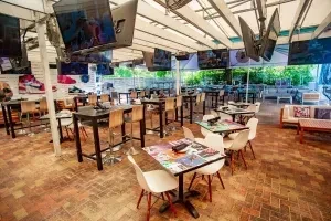 Open-air restaurant patio with overhead TVs, colorful tables, and lounge areas.