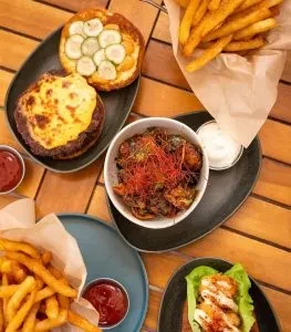 Wooden table with burger buns, fries, chicken, saucy stew, and sauces.