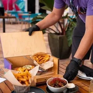 Gloved hands opening a wooden box of golden fries and food.