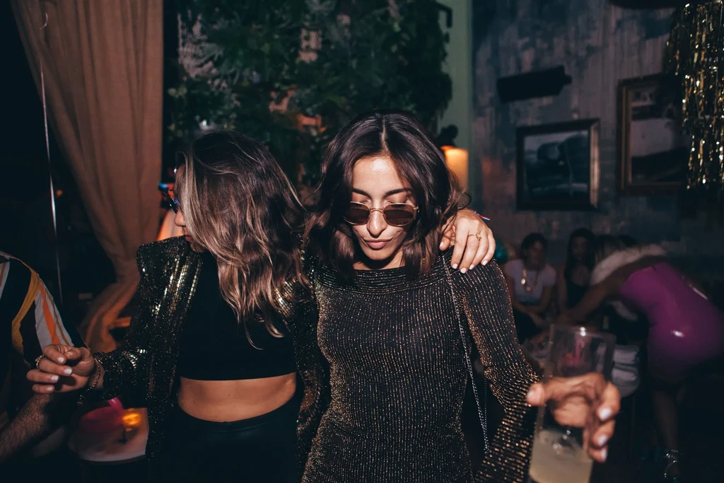 Two women in a dimly lit bar, one in sunglasses holding a drink.