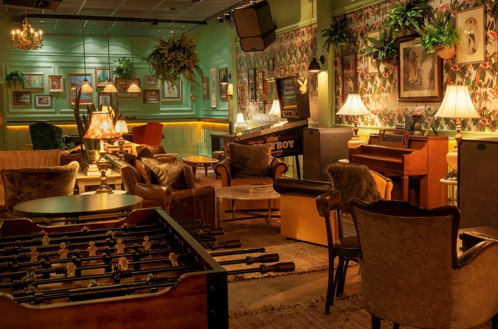 Eclectic lounge with foosball, pinball, plants, and tropical wallpaper.
