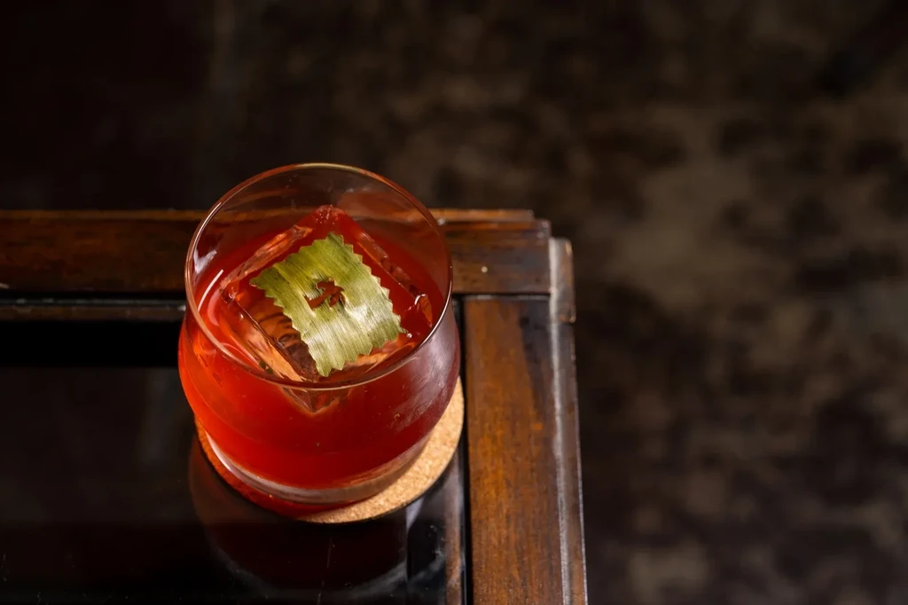 Reddish-amber cocktail with green leaf garnish on a square ice cube.