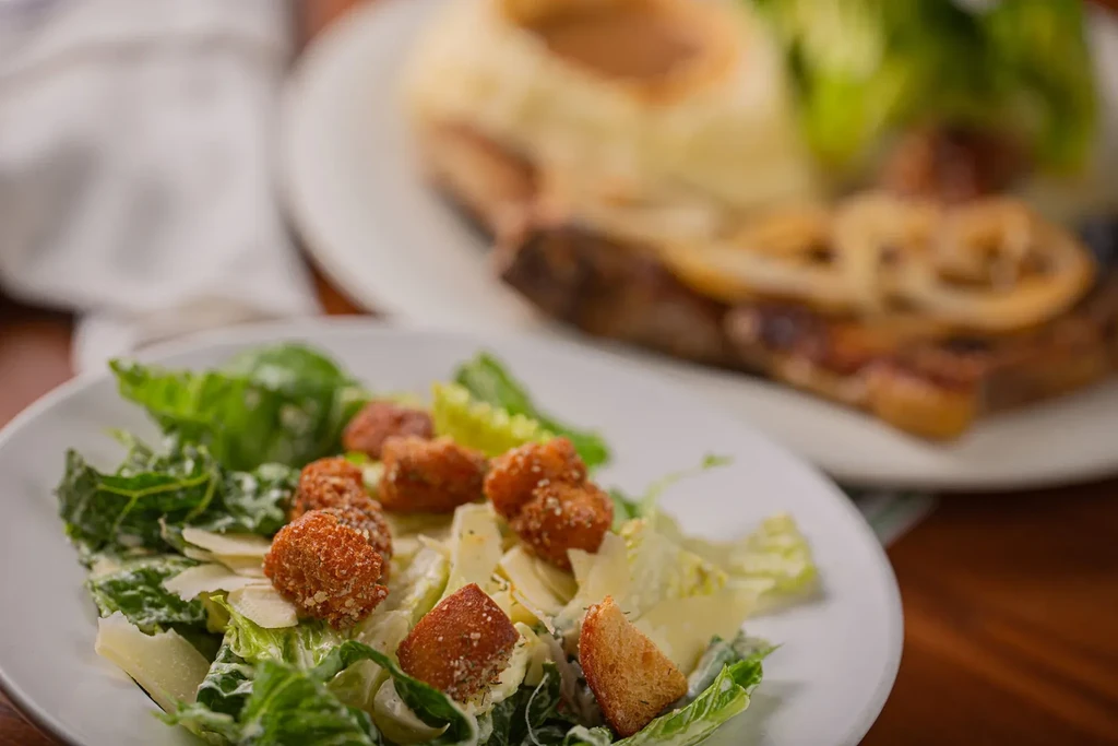 Caesar salad with fried items and croutons, blurred burger in background.