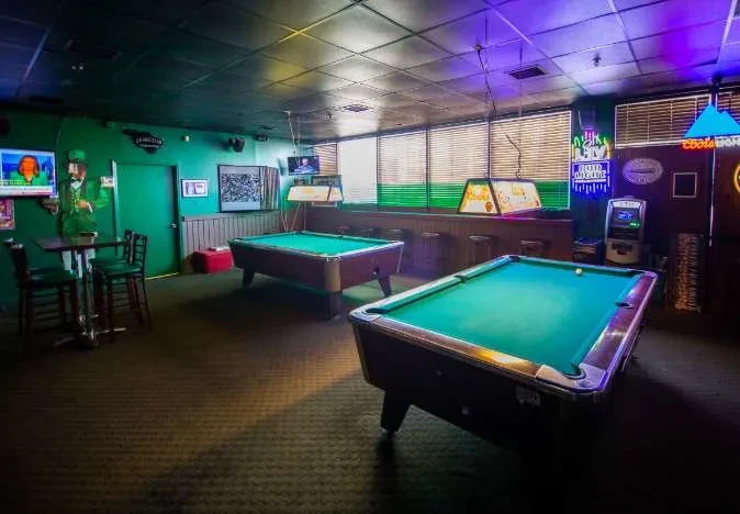 Dimly lit bar showing two pool tables, leprechaun, and neon signs.