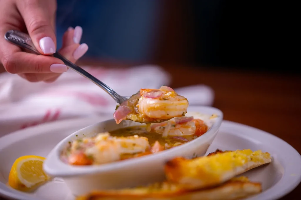 Hand spooning pinkish-orange shrimp with sauce, lemon, and bread.