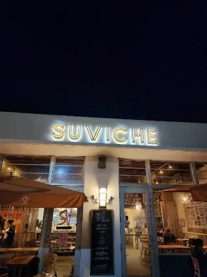 Illuminated SUVICHE restaurant storefront at night with indoor and outdoor diners.