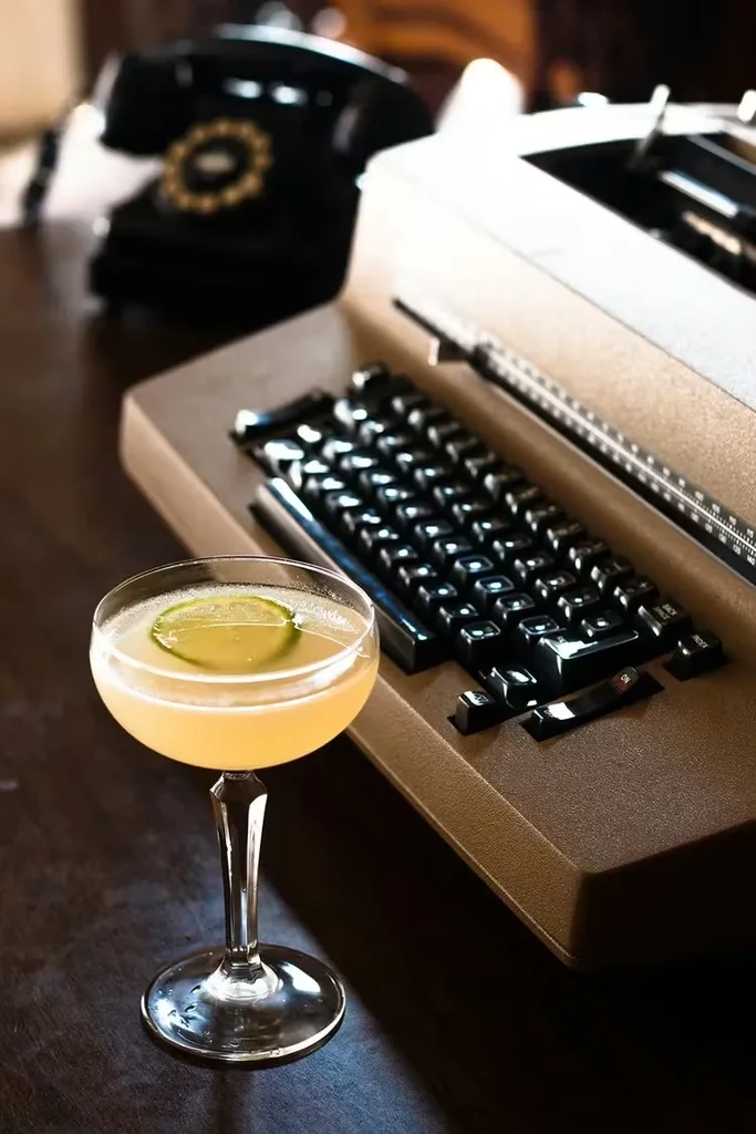 Pale yellow cocktail in coupe glass, vintage phone and typewriter.