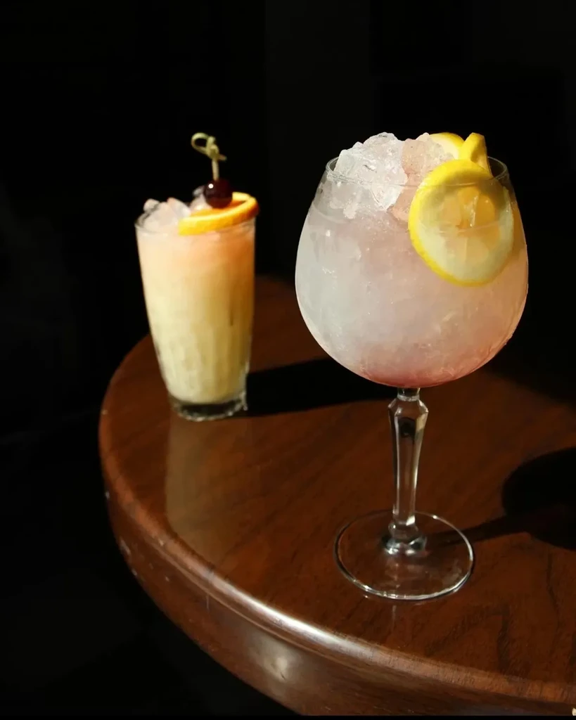 Two frosty cocktails: a pink goblet with lemon and an orange glass.