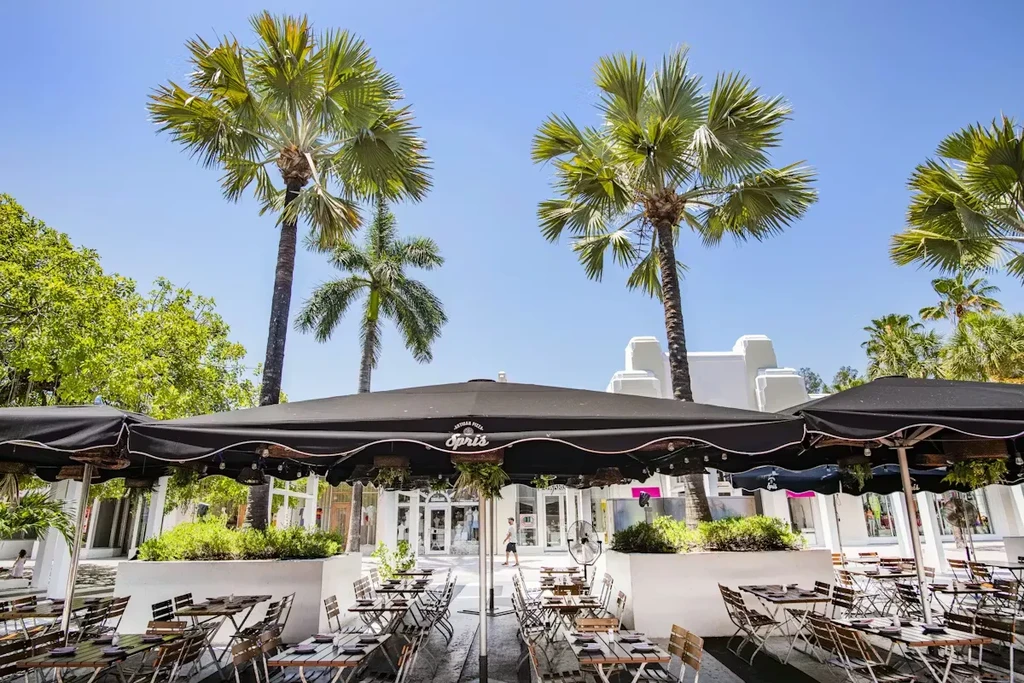 Sunny outdoor dining area with black umbrellas, one featuring "ARTISAN PIZZA Spris".